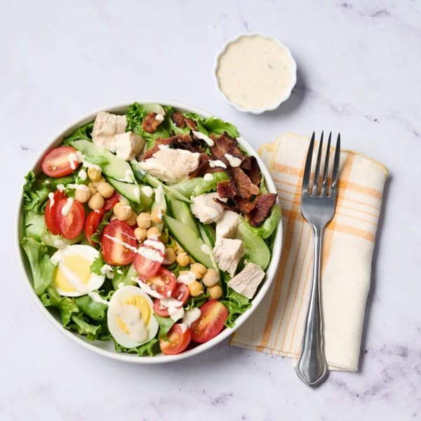Bowl of mixed greens with cherry tomatoes, cucumber, chickpeas, hard-boiled eggs, chicken, bacon bits, and a side of creamy dressing on a marble surface.