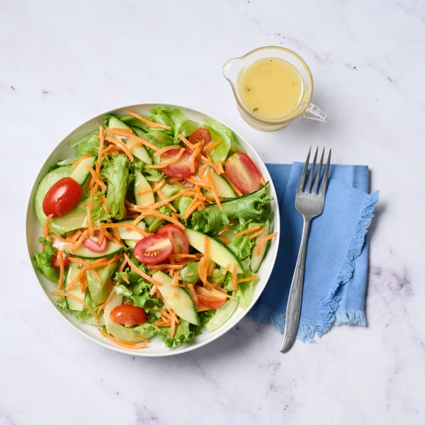 Bowl of mixed garden salad with lettuce, cucumber, cherry tomatoes, shredded carrot, and apple slices, with a small pitcher of vinaigrette nearby.