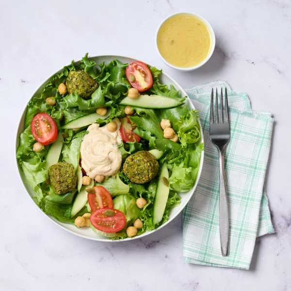Bowl of green salad with falafel bites, cucumber ribbons, cherry tomatoes, chickpeas, and a hummus dollop; dressing cup beside.