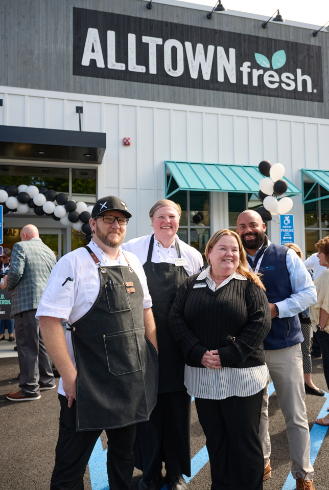 Group of four staff members posing outside Alltown Fresh during a storefront event, with balloons visible in the background.