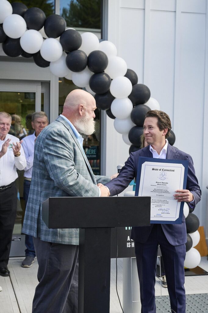Two men shake hands at an outdoor award ceremony, one holding a large certificate near a black-and-white balloon arch.